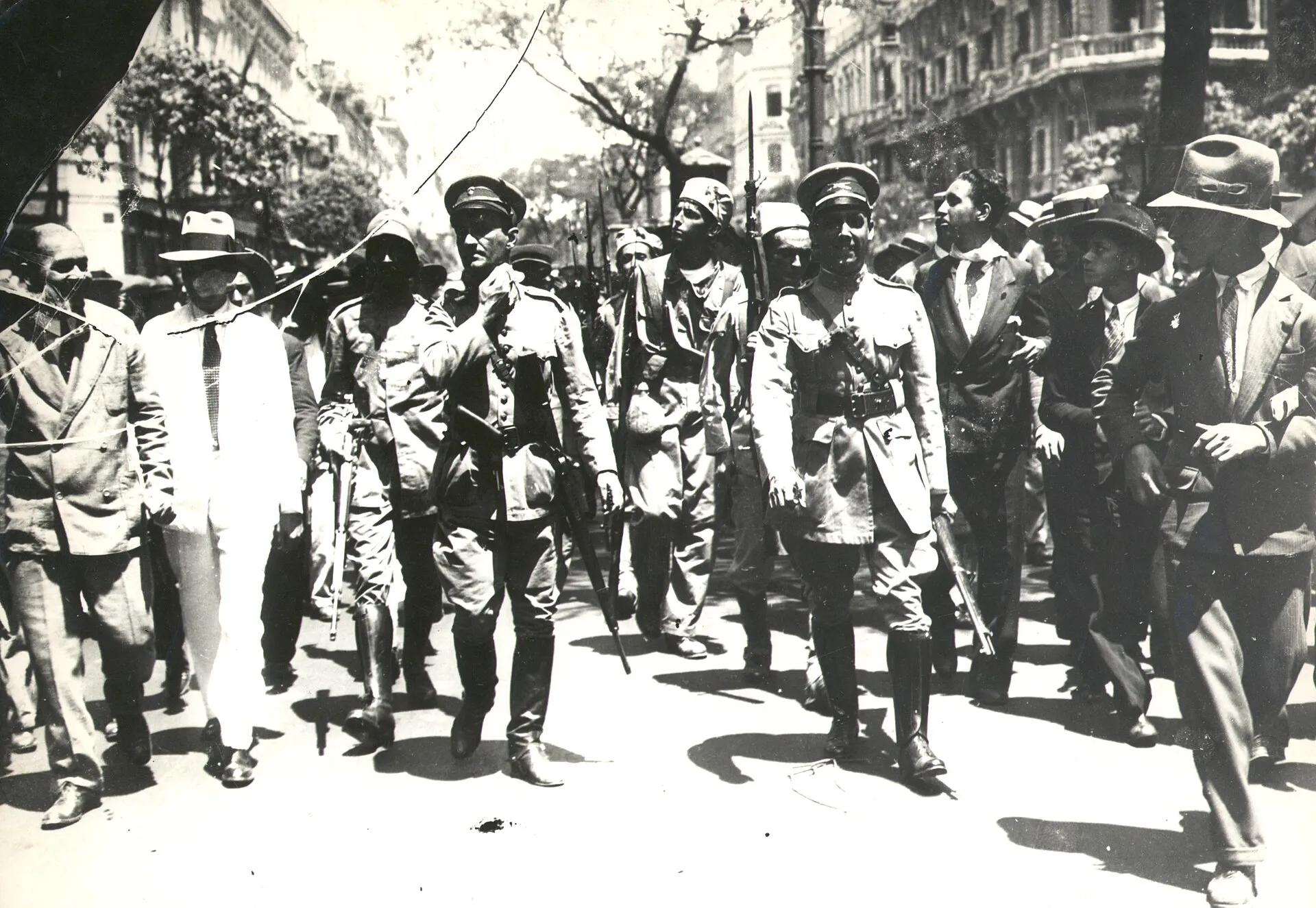 Foto em preto e branco de militares com fuzis e civis de terno e chapéu caminhando por uma avenida arborizada, em 1930
