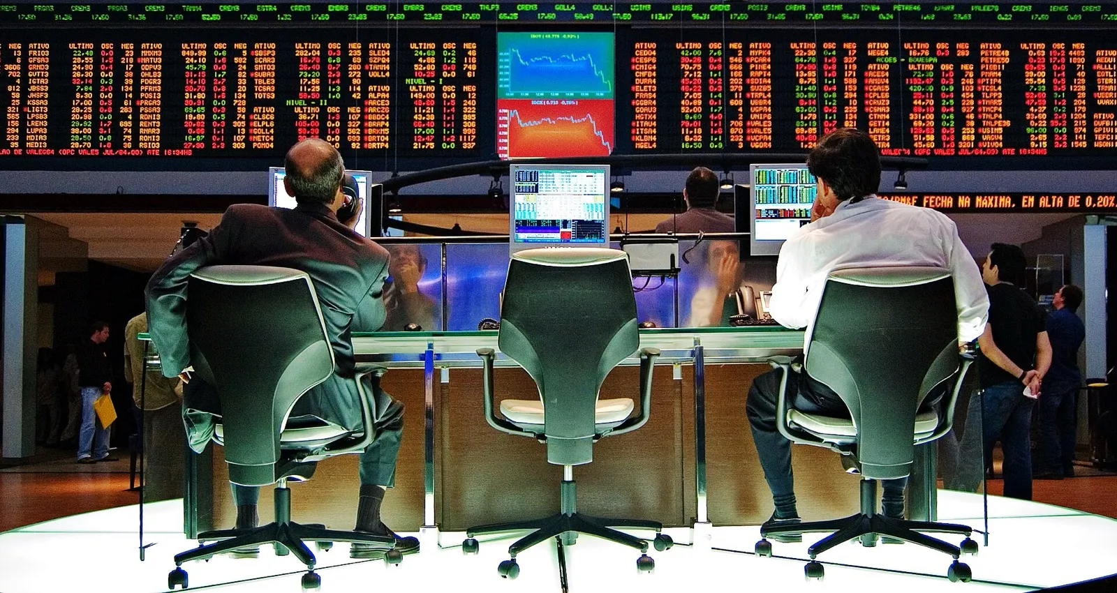 Dois operadores de costas diante de monitores, em frente a um painel de cotações na sala de negociação da Bovespa