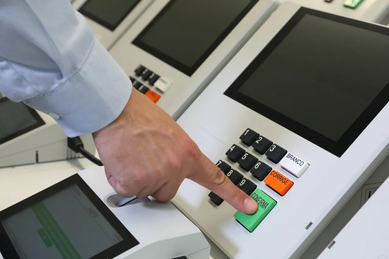 Mão digita em teclado branco de urna eletrônica, com teclas numeradas, teclas BRANCO, CORRIGE e CONFIRMA e pontos em braille nas teclas