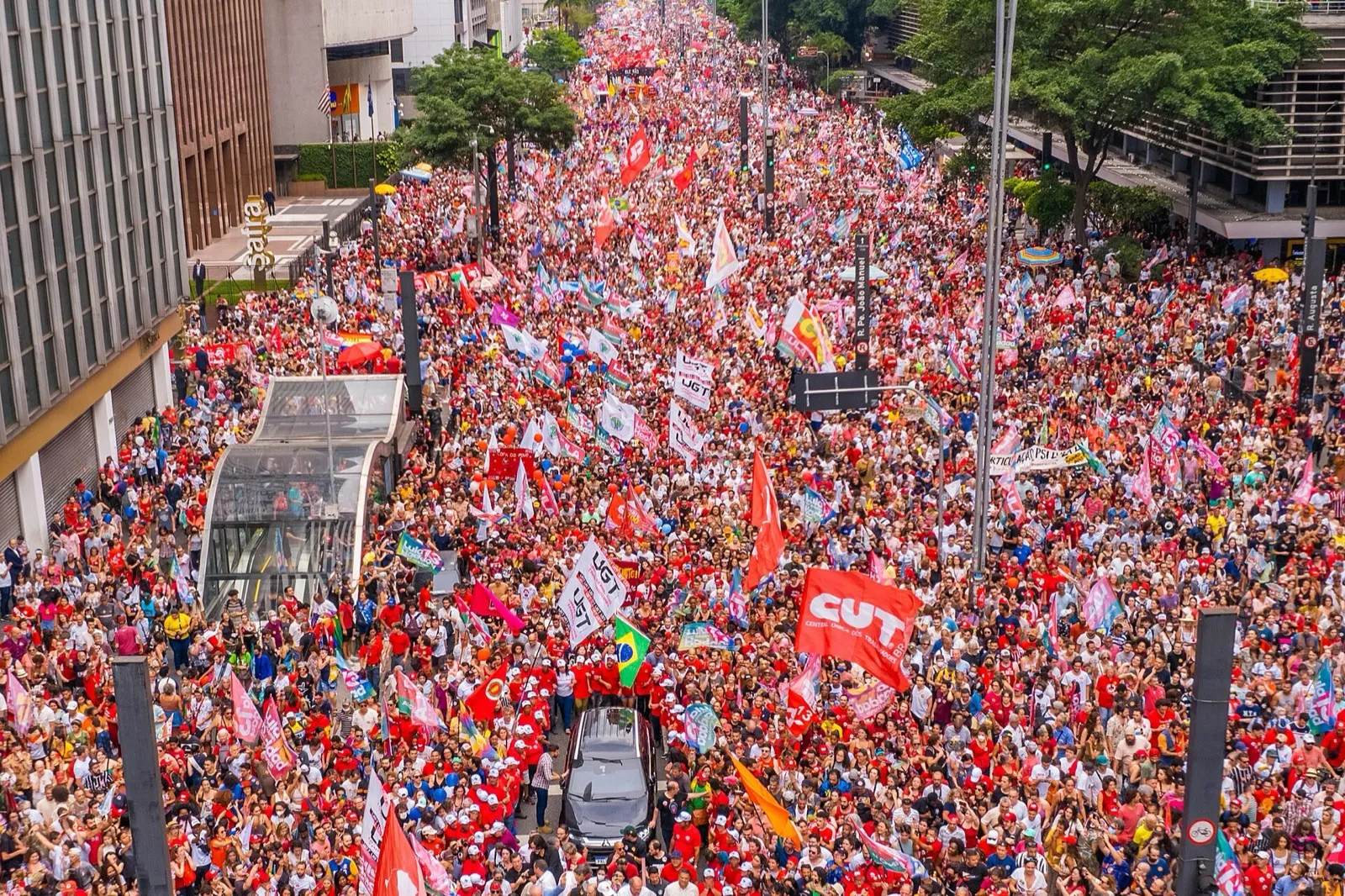 Vista aérea de uma multidão de camisetas vermelhas e bandeiras ocupando a Avenida Paulista, com prédios e árvores dos dois lados