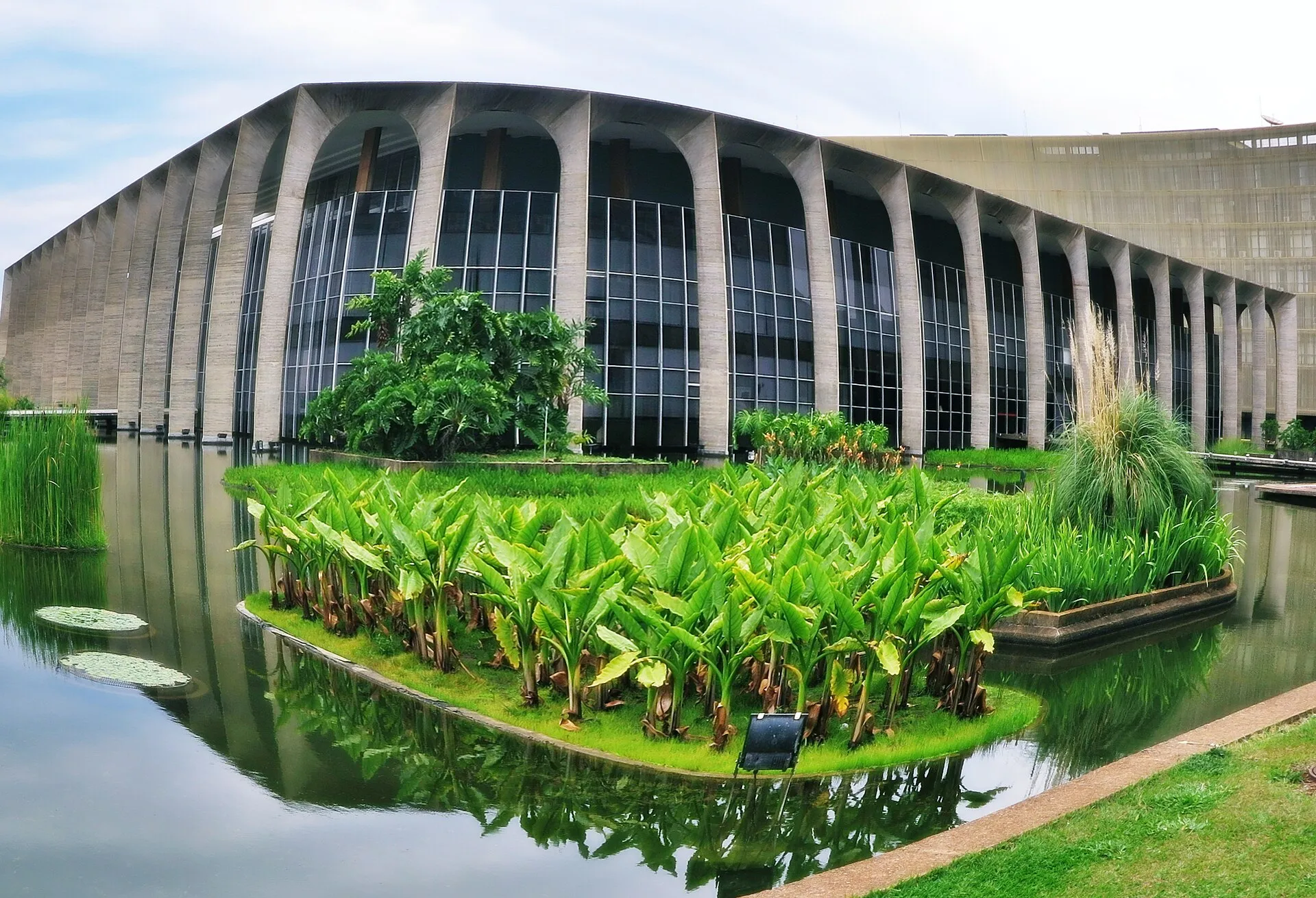 Fachada de arcos de concreto e vidro do Palácio do Itamaraty, em Brasília, refletida em espelho d'água com plantas aquáticas