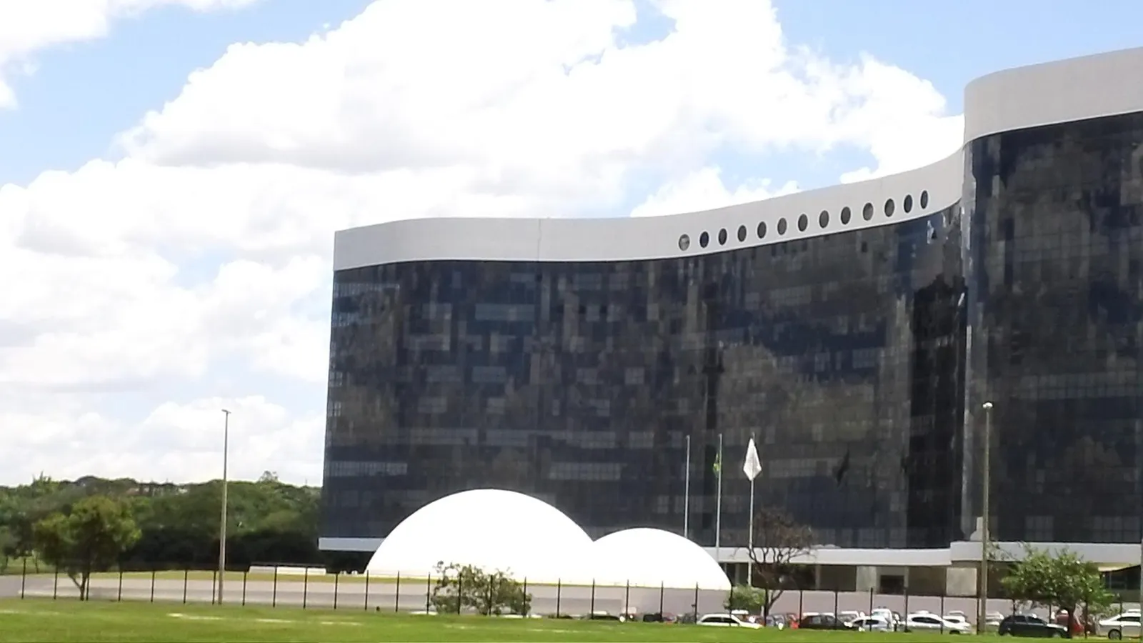 Fachada espelhada do edifício-sede do Tribunal Superior Eleitoral, em Brasília, com cúpulas brancas em primeiro plano e céu com nuvens