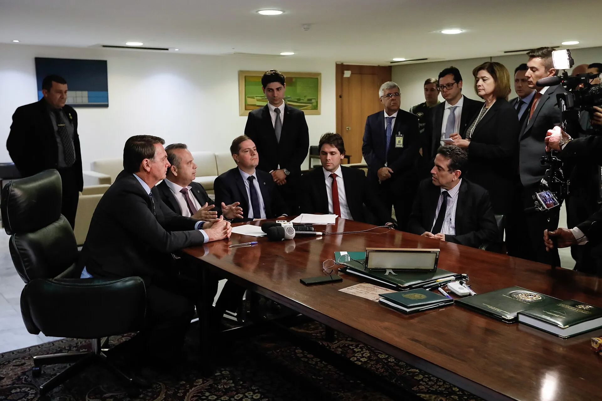 Homens de terno sentados e em pé ao redor de uma mesa de madeira no Palácio do Planalto, durante a assinatura de uma medida provisória, com cinegrafistas à direita