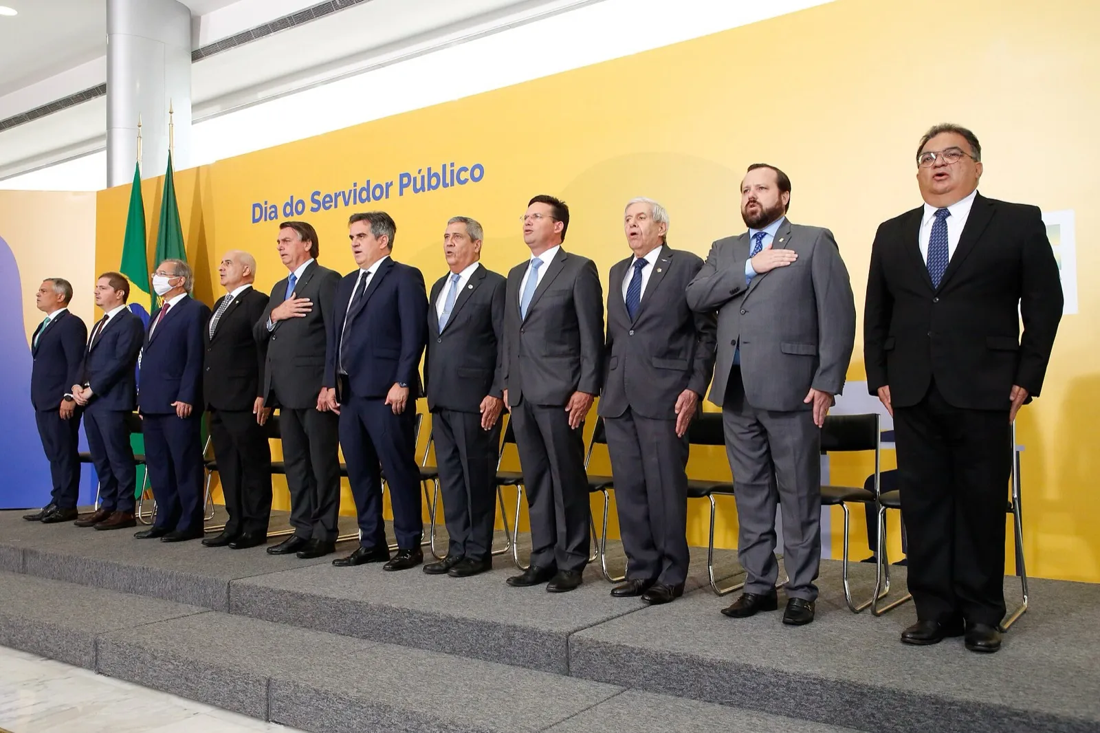 Autoridades de terno, em fila sobre um palco, diante de painel amarelo com a frase Dia do Servidor Público
