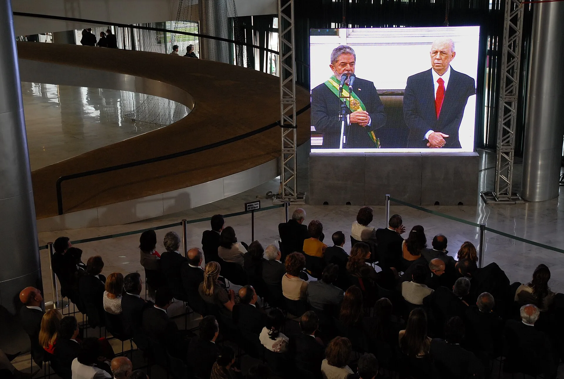 Telão no Palácio do Planalto mostra Lula com a faixa presidencial, diante de público sentado no saguão
