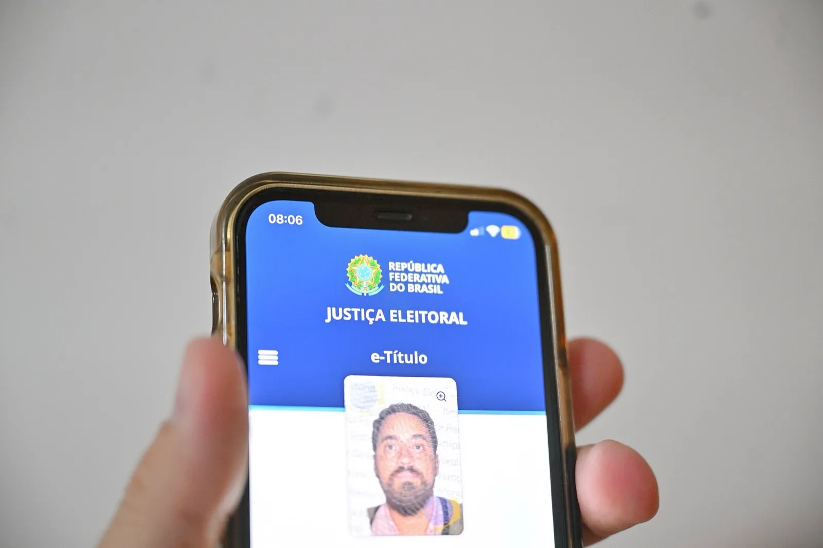 Mão segura celular com o aplicativo e-Título aberto, com o brasão da República e a palavra Justiça Eleitoral na tela