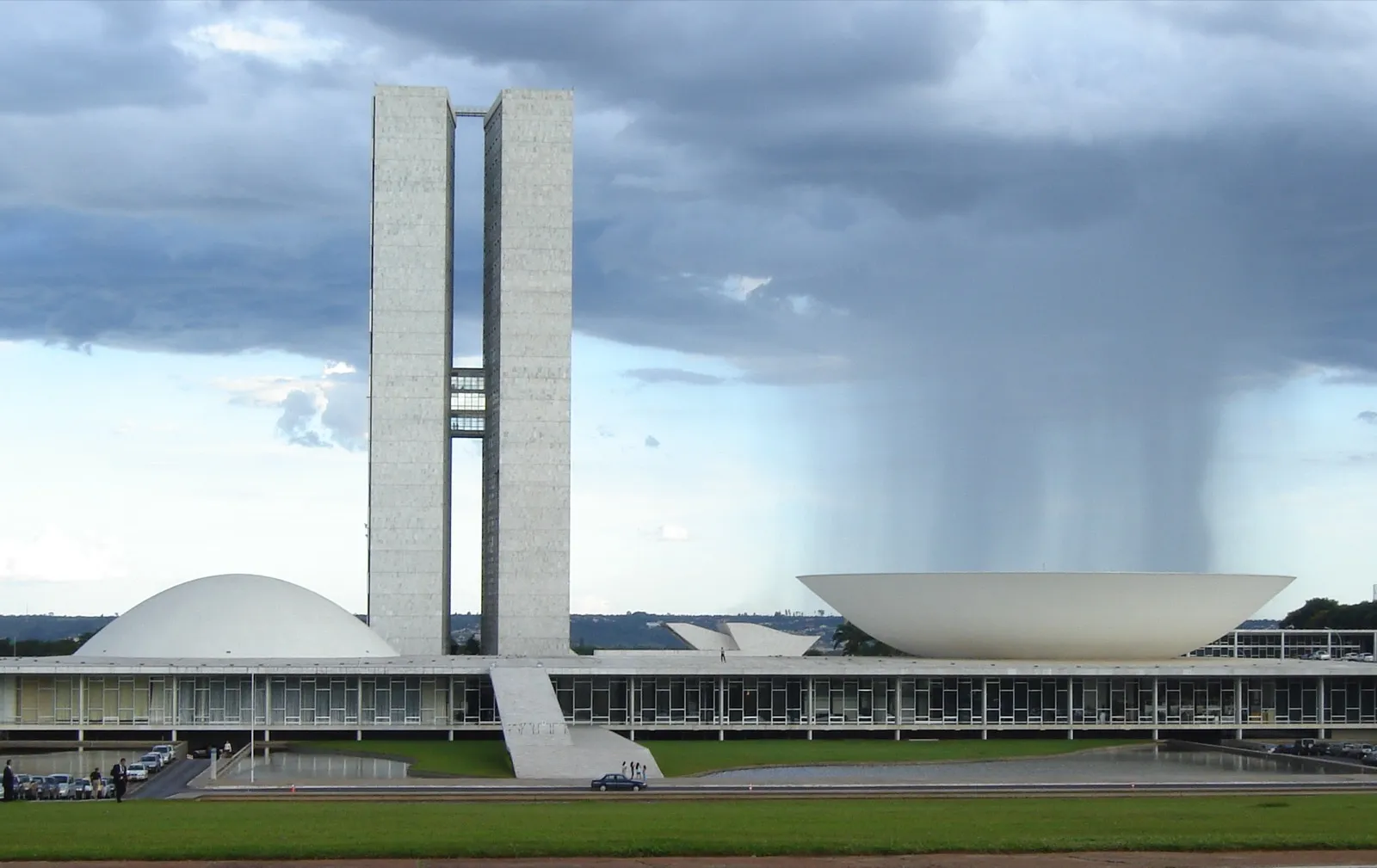 Prédio do Congresso Nacional em Brasília, com duas torres de concreto no centro, a cúpula do Senado à esquerda e a cúpula da Câmara à direita, sob céu nublado com chuva ao fundo
