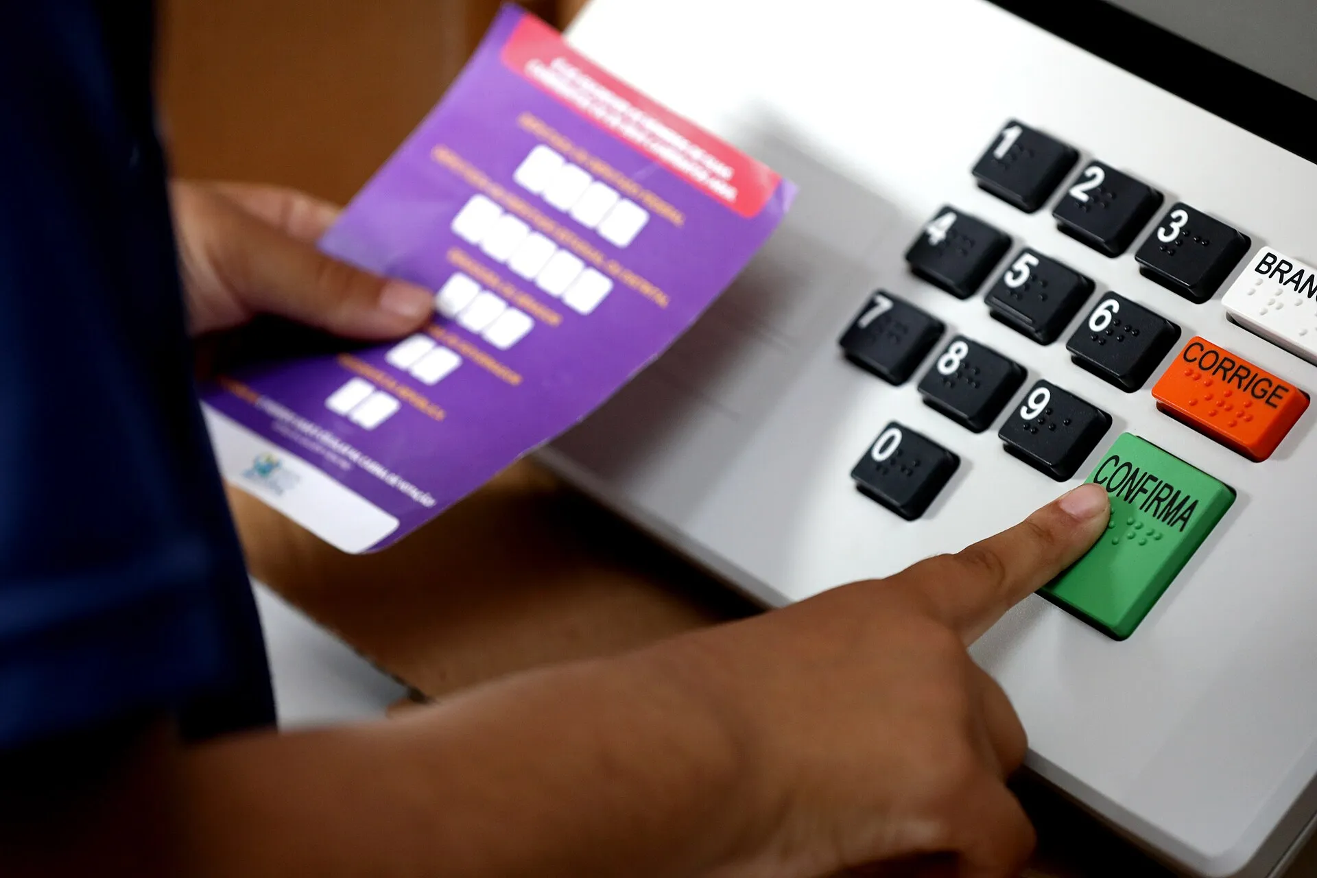 Mão aperta a tecla verde Confirma de uma urna eletrônica, enquanto outra mão segura uma cola de votação roxa