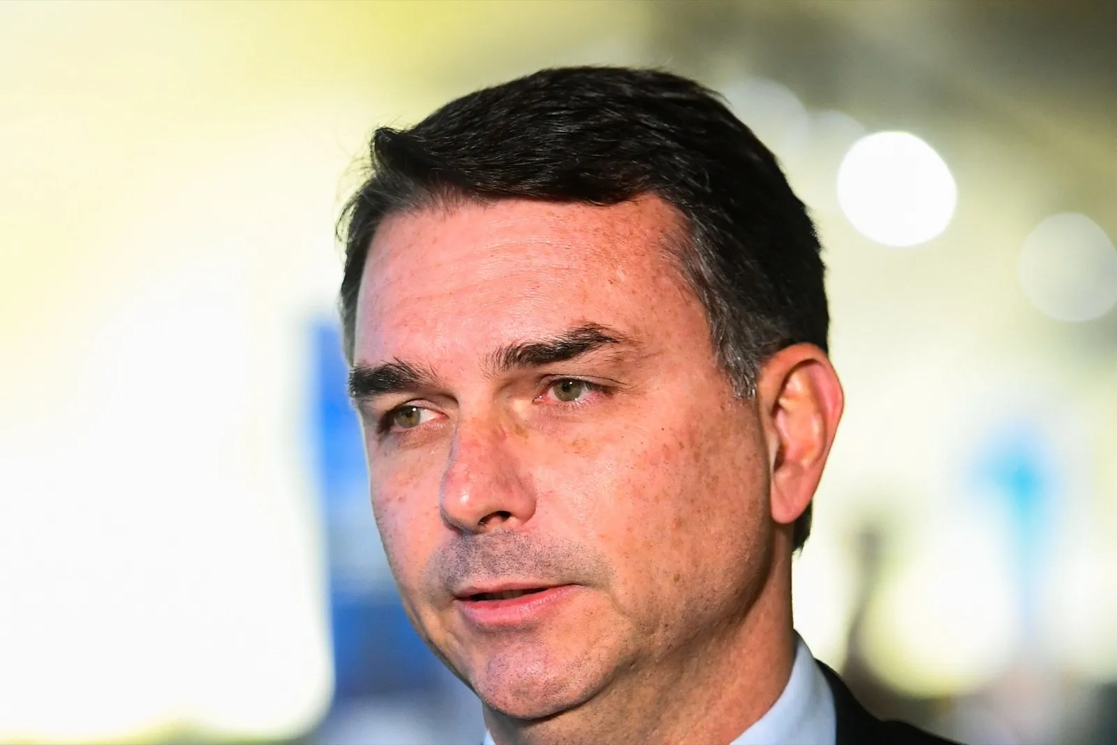 Senador Flávio Bolsonaro, de terno escuro, fala de perfil diante de um microfone da TV Senado