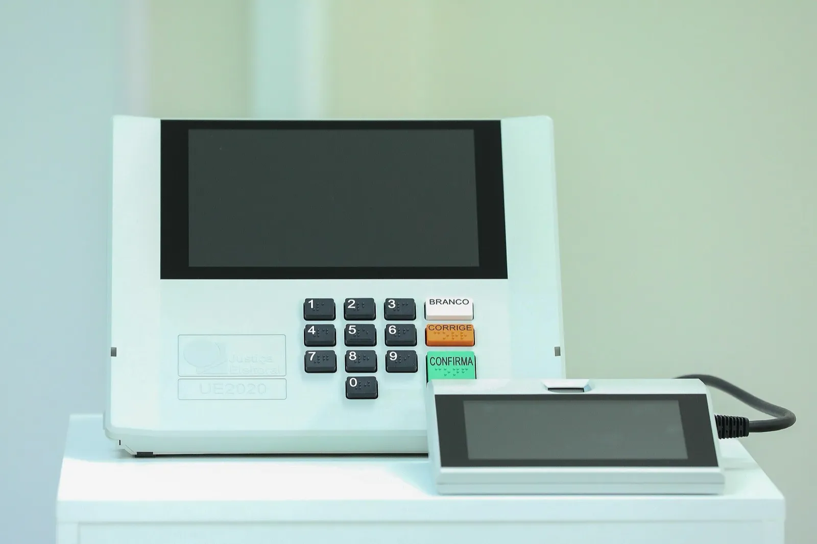 Urna eletrônica brasileira modelo UE2020, com tela, teclado numérico com braille e teclas BRANCO, CORRIGE e CONFIRMA, ao lado do terminal do mesário com leitor de digital