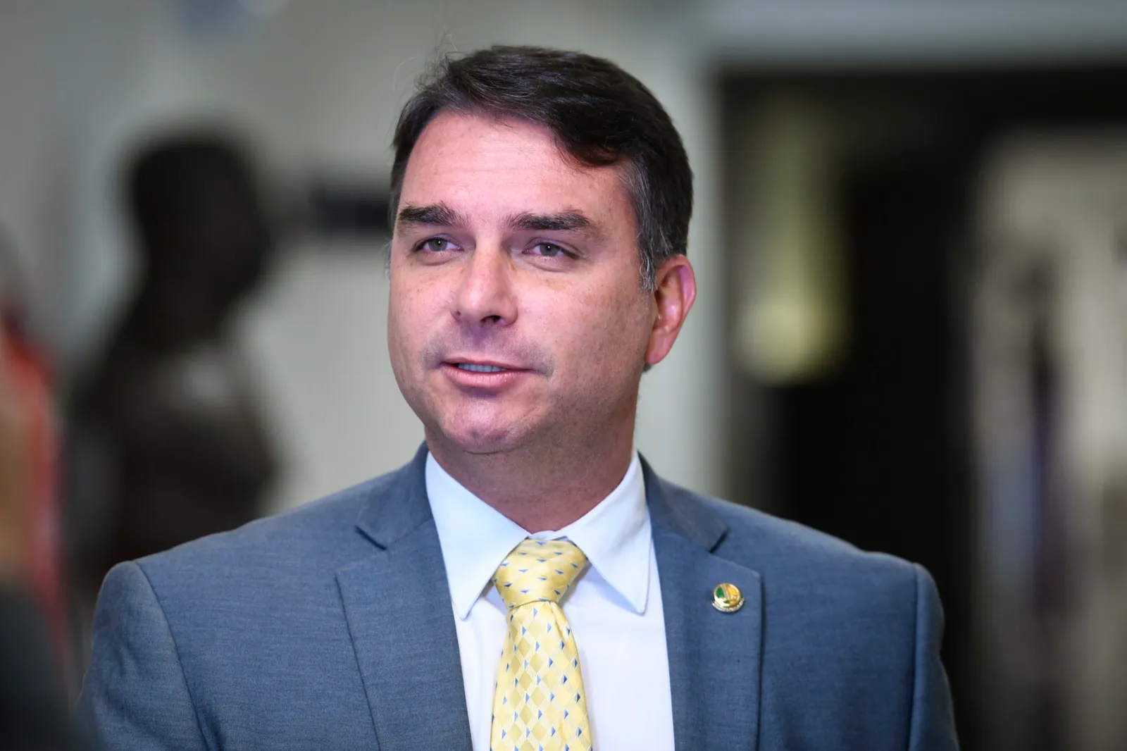 Flávio Bolsonaro, de terno cinza e gravata amarela, fala em entrevista em ambiente interno