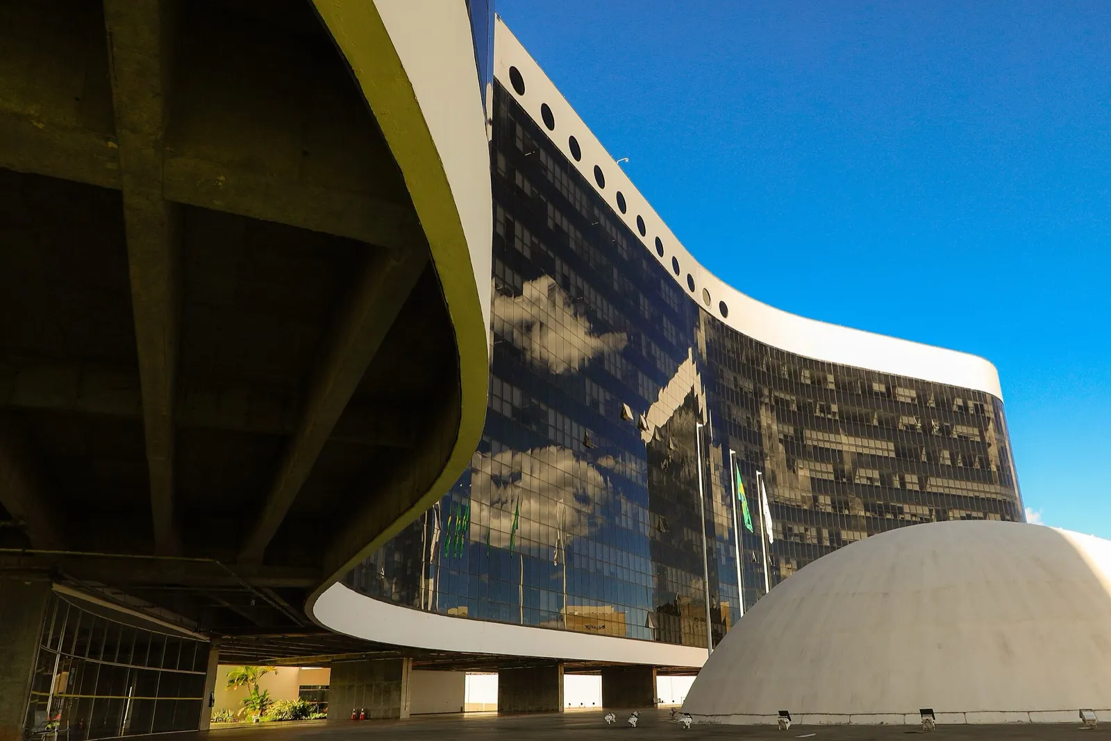 Fachada curva de vidro do edifício-sede do Tribunal Superior Eleitoral, com uma cúpula branca em primeiro plano e céu azul