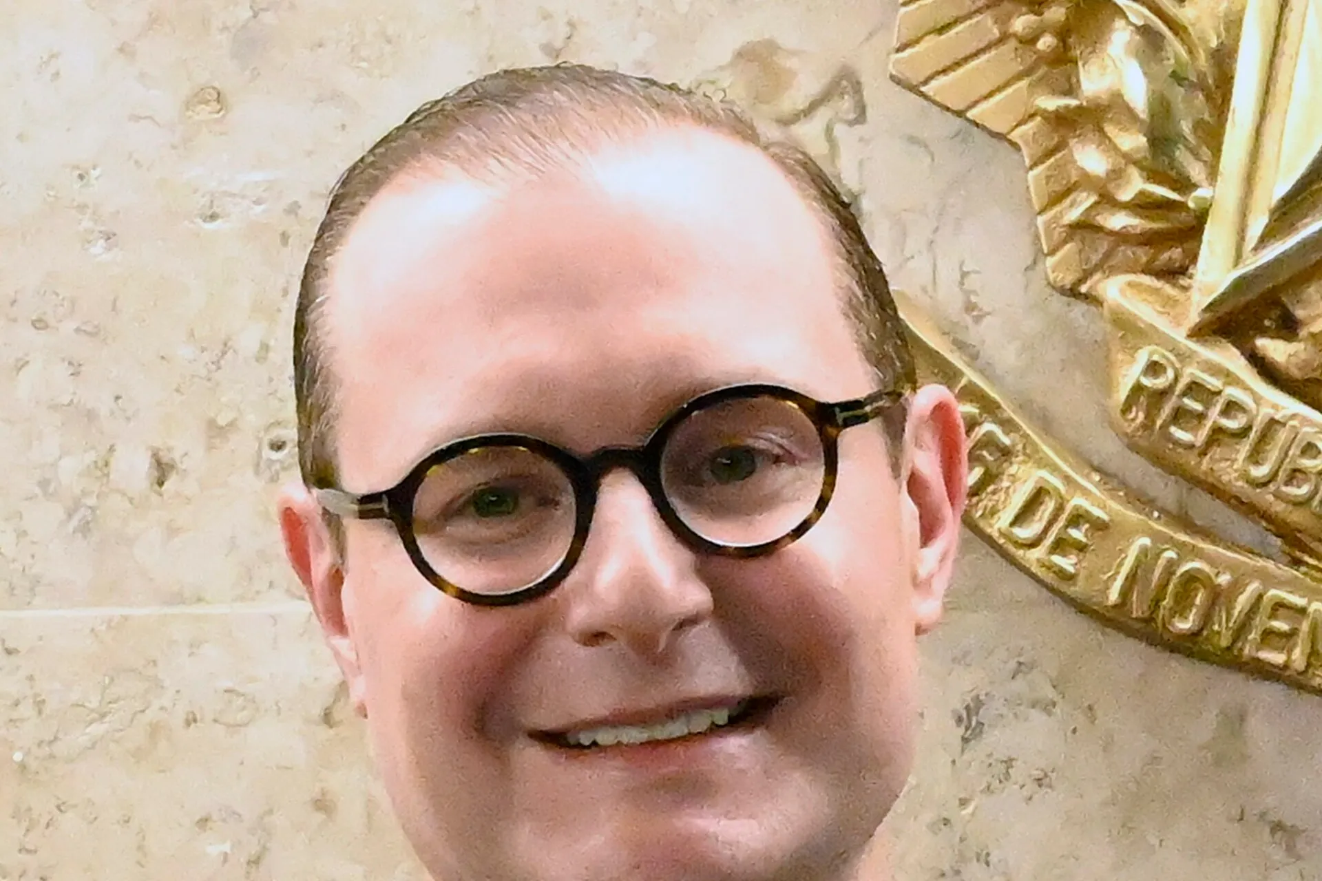 Retrato do ministro Cristiano Zanin, de toga preta, sorrindo, diante de uma parede de pedra clara com parte do brasão da República dourado